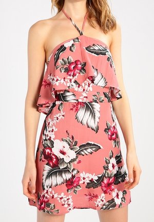 Femme portant une mini robe rose sans manches à col halter avec imprimé floral rouge, blanc et noir et un corsage à volants superposés.