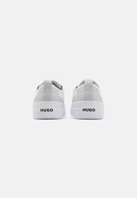 Une paire de baskets basses blanches avec des tirants gris et le logo "HUGO" à l'arrière des semelles en caoutchouc blanches, exposées côte à côte.