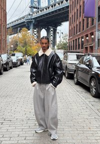 Black leather bomber jacket with white shearling collar, paired with gray wide-leg trousers and gray sneakers, set on a cobblestone street.