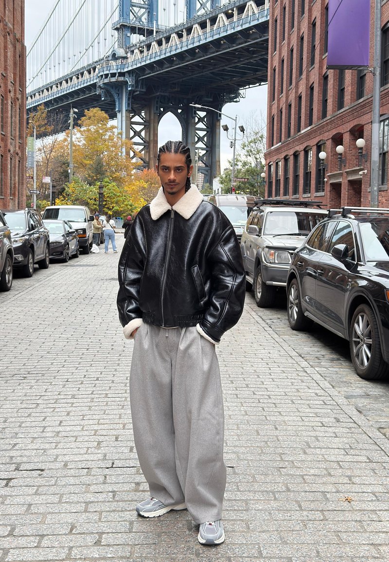 Black leather bomber jacket with white shearling collar, paired with gray wide-leg trousers and gray sneakers, set on a cobblestone street.