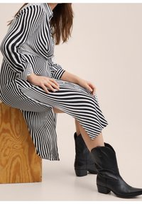 Femme assise sur une caisse en bois portant une robe à rayures noires et blanches et des bottes en cuir noir mi-mollet, sur fond neutre.