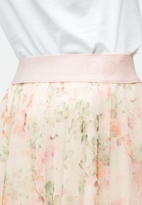 Κοντινό πλάνο ενός ελαφρού φλοράλ τούλινης φούστας με φαρδύ ανοιχτό ροζ ζωνάρι μέσης που διαθέτει διακριτικό ανάγλυφο κείμενο, φορεμένη με λευκό τοπ.