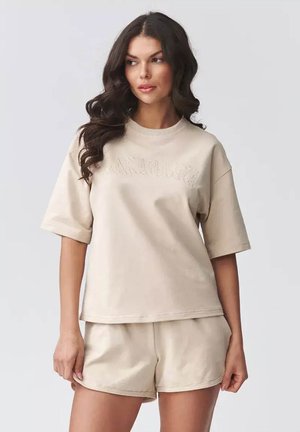 Kobieta z długimi kręconymi ciemnymi włosami, ubrana w beżowy, oversize T-shirt i pasujące szorty, stoi na jasnym, jednolitym tle.