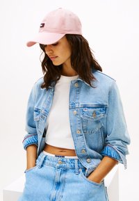 Gorra rosa claro con logo, chaqueta de mezclilla con lavado azul, top corto blanco, pantalones cortos de mezclilla azul de cintura alta; atuendo casual con texturas en capas.