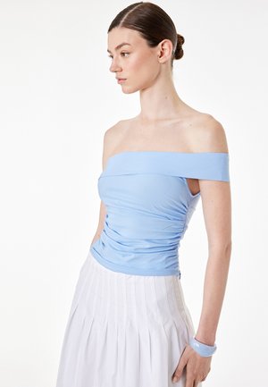Joven mujer con el cabello recogido en un moño que lleva una blusa azul claro sin hombros, falda blanca plisada y una pulsera azul a juego, mirando hacia la izquierda sobre fondo blanco.