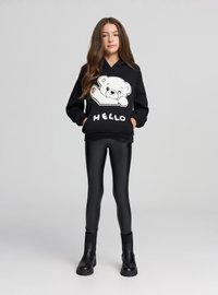 Czarna bluza z kapturem z białą grafiką niedźwiedzia i napisem "HELLO", połączona z czarnymi błyszczącymi legginsami i czarnymi botkami za kostkę.