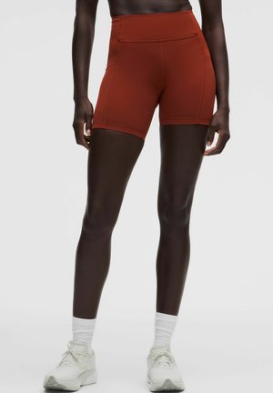 Person iført rustfarvede, højtaljede sportsshorts, hvide crew sokker og hvide sneakers, stående mod en ensartet grå baggrund.