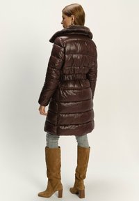 Hnědý péřový dlouhý kabát s načechraným kožešinovým límcem. Má páskový pas a lesklou texturu, doplněný koženými vysokými botami po kolena.