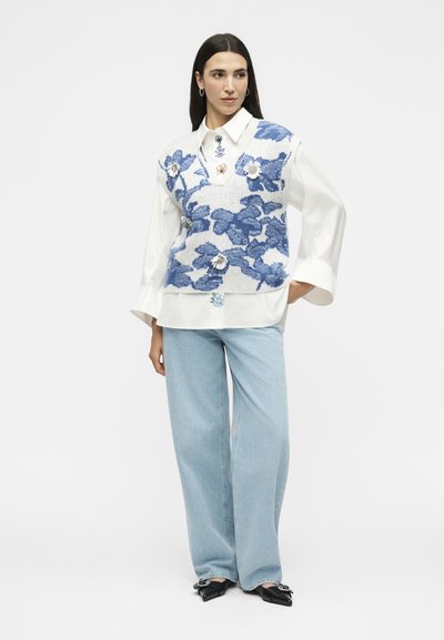 Femme portant une chemise blanche avec une broderie florale bleue, un gilet en maille bleu et blanc à motifs floraux, un jean large bleu clair et des chaussures noires à bout pointu.