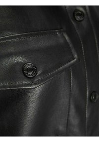 Musta nahkatakki, jossa on rintatasku mustalla napilla, teksturoitu pinta ja näkyvät saumat reunoilla.