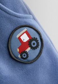 Modrá flísová tkanina s kruhovou vyšitou nášivkou s návrhom červeného traktora, čiernymi kolesami a kontrastným svetlomodrým pozadím.