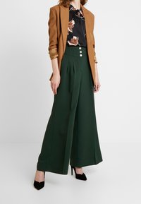 Brun blazer over en sort blomsterbluse, parret med grønne brede bukser med tre frontknapper og en glat tekstur. Sorte hæle fuldender looket.