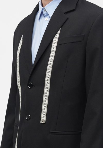 Personne portant un blazer noir et une chemise bleu clair avec un ruban à mesurer blanc suspendu autour du cou sur fond blanc.