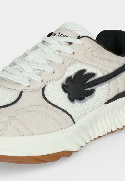 Λευκό και μπεζ αθλητικό παπούτσι με μαύρο σχέδιο φύλλου σφενδάμου, υφασμάτινο διχτυωτό υλικό, λευκά κορδόνια και παχύ λευκό πέλμα με καφέ πέλμα.