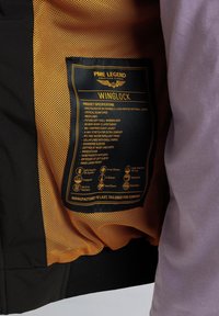 Schwarz-orange Softshell-Jacke mit Netzfutter, Produktetikett mit Details zu Materialien und Funktionen, einschließlich wasserabweisendem und dehnbarem Stoff.