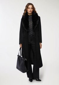 Lungo cappotto nero con colletto in pelliccia sintetica, abbinato a una maglietta nera a pois e pantaloni neri su misura. La modella porta una borsa nera.