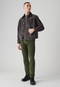 Chaqueta de mezclilla gris oscuro con cuello de piel sintética negra, pantalones de pana verde y zapatos negros con cordones, sobre un fondo gris claro.
