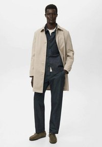 Trench coat beige, camicia di jeans scura e pantaloni blu scuro. Scarpe marroni. Texture lisce, dettagli dei bottoni e abbigliamento a strati visibili.