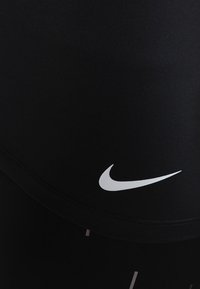 Zwarte atletische leggings gemaakt van rekbaar materiaal met een wit Nike-logo op de heup en subtiele patroondetails op het onderste deel van het been.