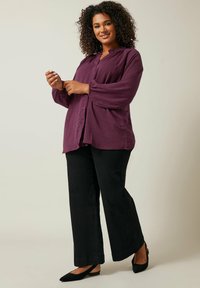 Blusa viola con bottoni, realizzata in un tessuto leggermente testurizzato e con maniche arricciate, abbinata a pantaloni neri a gamba larga e scarpe flat nere.