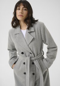 Cappotto doppiopetto grigio in misto lana con cintura, grandi bottoni neri, colletto a scannellature e tasche laterali, che mette in mostra una texture morbida.