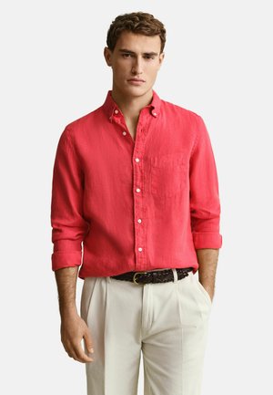 Junger Mann, der ein hellrotes Hemd mit hochgekrempelten Ärmeln und weiße Hosen mit einem geflochtenen schwarzen Gürtel trägt, steht mit einer Hand in der Tasche.