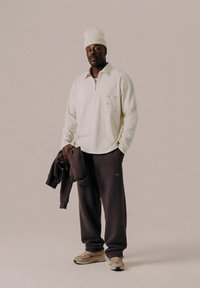 Man wearing a white knit hat, cream long-sleeve shirt, dark sweatpants, beige sneakers, and holding a dark sweatshirt standing against a plain background.