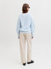 Persoon in lichtblauwe gebreide trui, beige wijde broek, zwarte schoenen en zonnebril, staand met de rug naar voren op witte achtergrond.