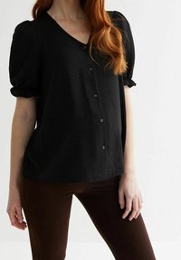 Haut noir à manches courtes en V avec des manches bouffantes, présentant une fermeture à boutons sur le devant et une coupe décontractée, associé à un legging marron foncé.