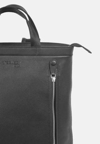 Svart läder tote-väska med texturerad yta, topphandtag och en sidoficka med dragkedja. Har en logotyp präglad på framsidan.