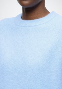 Světle modrý svetr z měkkého materiálu, s žebrovaným kulatým výstřihem a raglánovými rukávy. Hladká textura s minimálním zdobením.