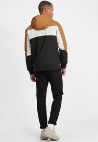 Mann i en fargeblokk jakke med brune, hvite og svarte seksjoner, svarte jeans, hvite sokker og beige joggesko, som står med ryggen til.