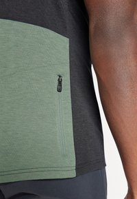 Camisa de manga corta con un panel frontal texturizado en verde y parte trasera negra, con un bolsillo con cremallera en el lado izquierdo para almacenamiento.
