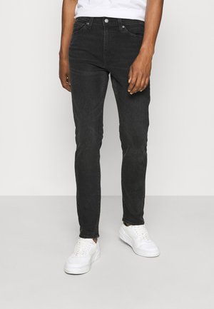 Homme portant un jean noir slim et des baskets blanches, debout devant un fond blanc uni.