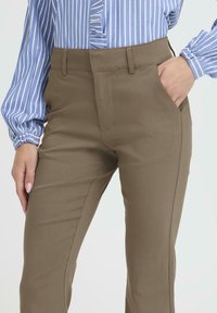 Pantalones marrones, ajustados y a medida, con un corte slim, que cuentan con bolsillos laterales y una textura suave. Llevados con una blusa a rayas azul y blanca.