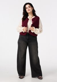 Gilet en polaire bordeaux porté sur un chemisier crème à manches volantes, associé à un jean noir à jambes larges et des talons à motifs.