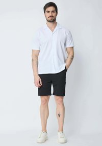Chemise blanche à manches courtes avec col, associée à un short noir et des baskets blanches. Comprend des tatouages visibles sur l'avant-bras.