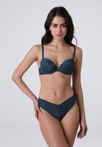Donna con lunghi capelli scuri che indossa un set coordinato di reggiseno e mutandine in pizzo teal scuro, in piedi su uno sfondo grigio chiaro uniforme.