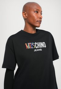 Juodas marškinėliai su apvalia iškirpte ir trumpomis rankovėmis, puošti spalvinga, žaisminga užrašu "MOSCHINO JEANS". Tekstūruota medžiaga ir laisvas fasonas.