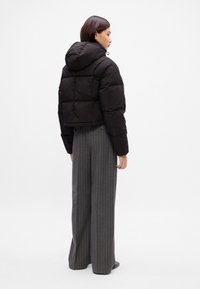 Casaco puffer preto cropped com capuz, apresentando painéis cosidos. Modelo veste calças largas cinza com riscas.