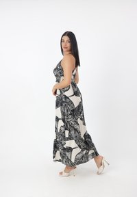 Robe maxi à imprimé feuillage noir et crème, avec des bretelles fines et un corsage ajusté, s'évasant au bas, portée avec des sandales à talons blanches.