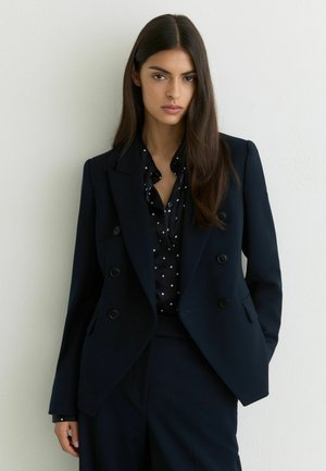 Kobieta o długich ciemnych włosach, ubrana w czarną marynarkę dwurzędową na czarnej bluzce w białe grochy, stojąca przy białej ścianie.