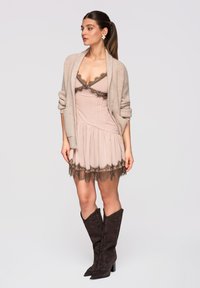 Femme debout portant une robe nuisette beige avec une bordure en dentelle foncée, un cardigan beige aux manches retroussées, et des bottes hautes marron foncé.