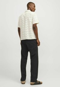 Camisa de manga corta en beige claro con patrón de ojales florales, combinada con pantalones gris oscuro y sandalias marrones. Vista trasera del modelo.