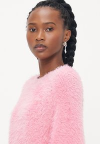 Roze donzige trui met een gestructureerde afwerking, met een klassieke ronde halslijn. Het model heeft gevlochten haar en draagt zilveren ringoorbellen.