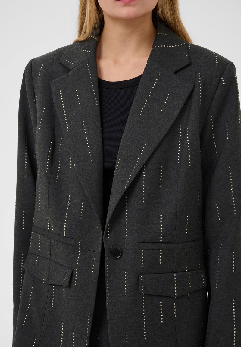 Blazer nero con motivo a pois argentati, dotato di revers, due tasche frontali e chiusura a un bottone. Tessuto morbido.