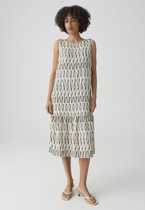 Vrouw met krullend haar die een mouwloze witte midijurk draagt met een zwart golvend streeppatroon en beige sandalen met hakken, staand op een witte achtergrond.