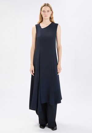 Donna in piedi che indossa un abito nero asimmetrico senza maniche sopra pantaloni neri a gamba larga e scarpe nere su sfondo bianco.