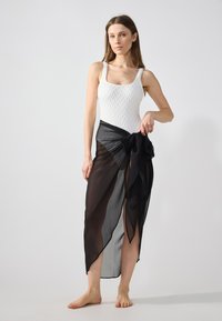 Jupe transparente noire avec un détail de nœud, associée à un body ajusté blanc en texture. Le mannequin se tient pieds nus sur un fond clair.