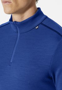 Modrý pulovr s vysokým límcem a polovičním zipem. Vyrobený z texturované látky, zdobený kontrastními švy a malým logem na límci.